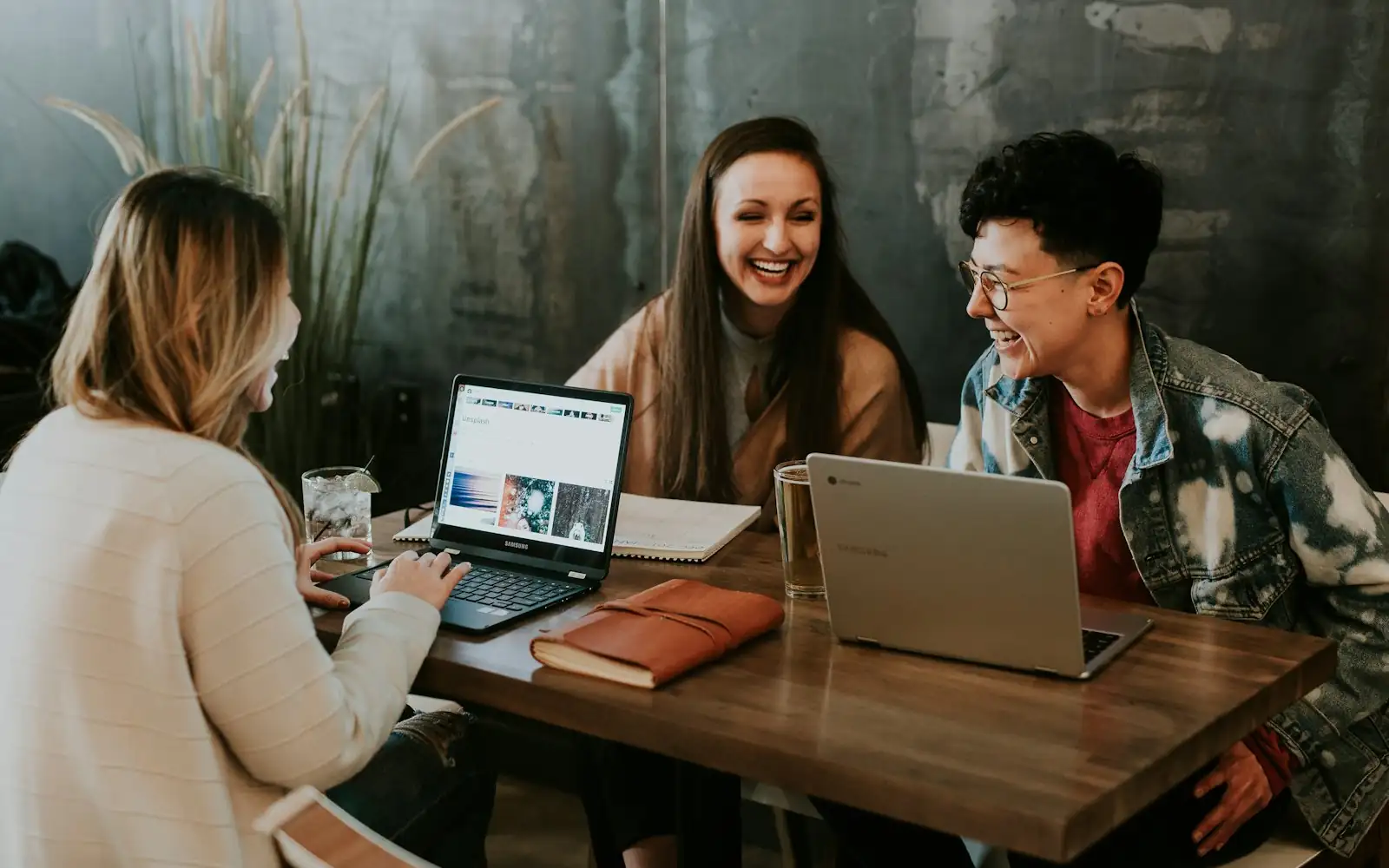 Three colleagues laughing around laptops at a wooden table
